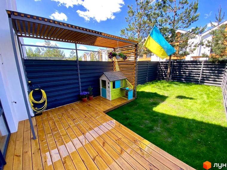 Вигляд подвір'я Котеджного містечка Prydesennya Eco Village з терасою, дитячим будиночком та газоном. На задньому плані видно паркан.