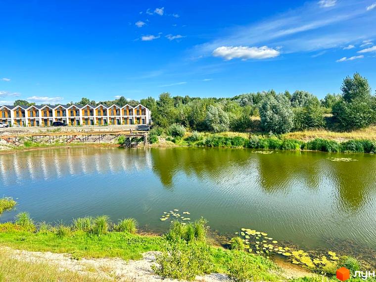 Вигляд на Котеджне містечко Prydesennya Eco Village біля озера. На передньому плані зелена трава та вода, а на задньому будинки.