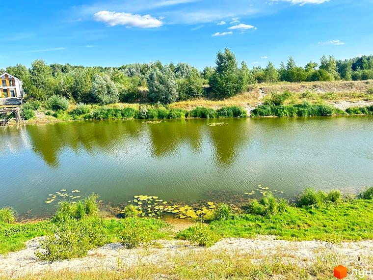 Вигляд на будинок Котеджного містечка Prydesennya Eco Village з річки. Навколо зелені дерева та трава.