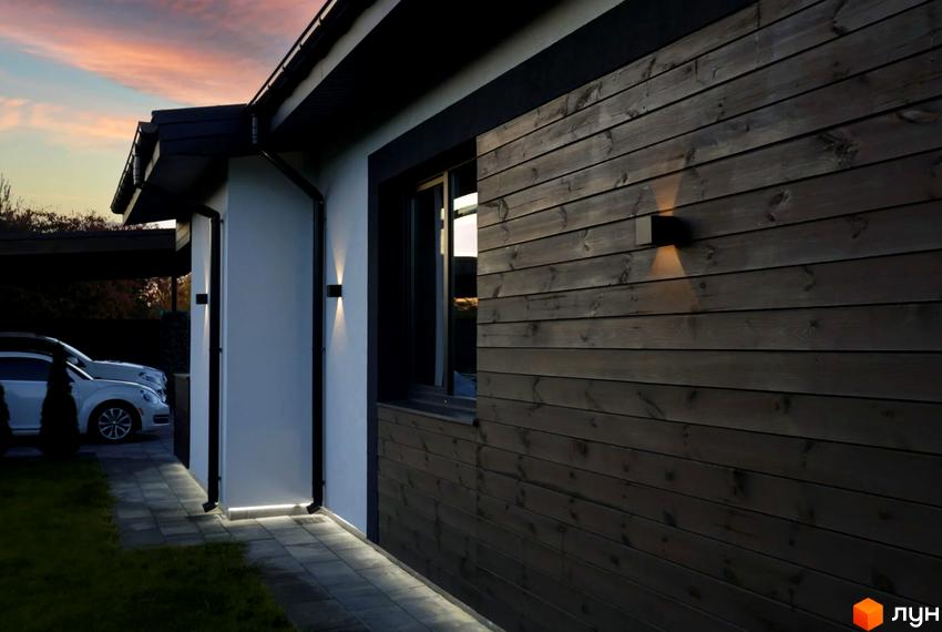Зовнішній вигляд будинку з дерев'яним фасадом та підсвіткою. Прибудинкова територія з доріжкою та автомобілями біля котеджного містечка КМ New Village.