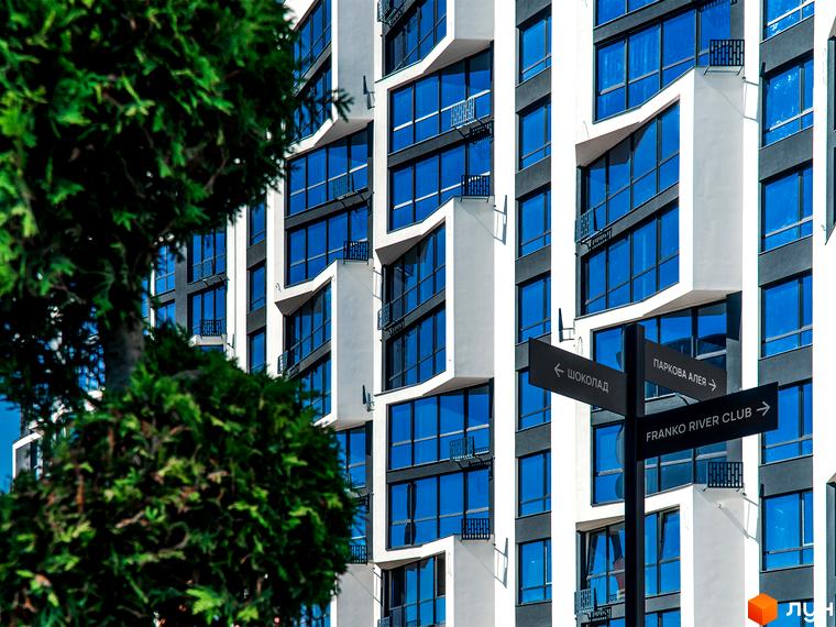 Зовнішній вигляд будинку в Житловому комплексі Паркова Алея з великими вікнами. На передньому плані зелені дерева та покажчик напрямків.