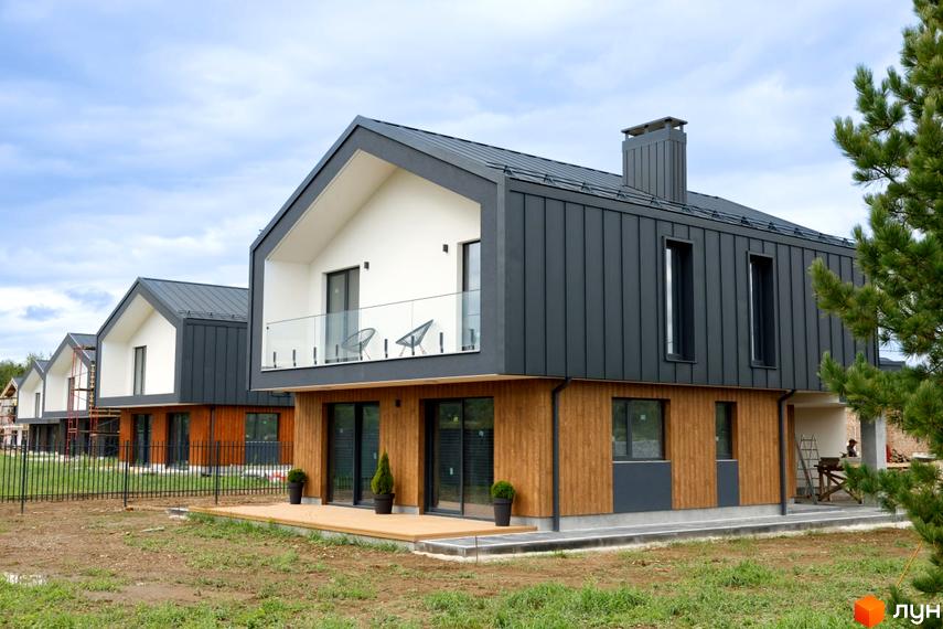 Будинки Котеджного містечка OZON Village мають темно-сірі дахи та фасади з металу, білі фронтони та дерев'яну обшивку. Навколо будинків видно ділянки з травою та огорожу.