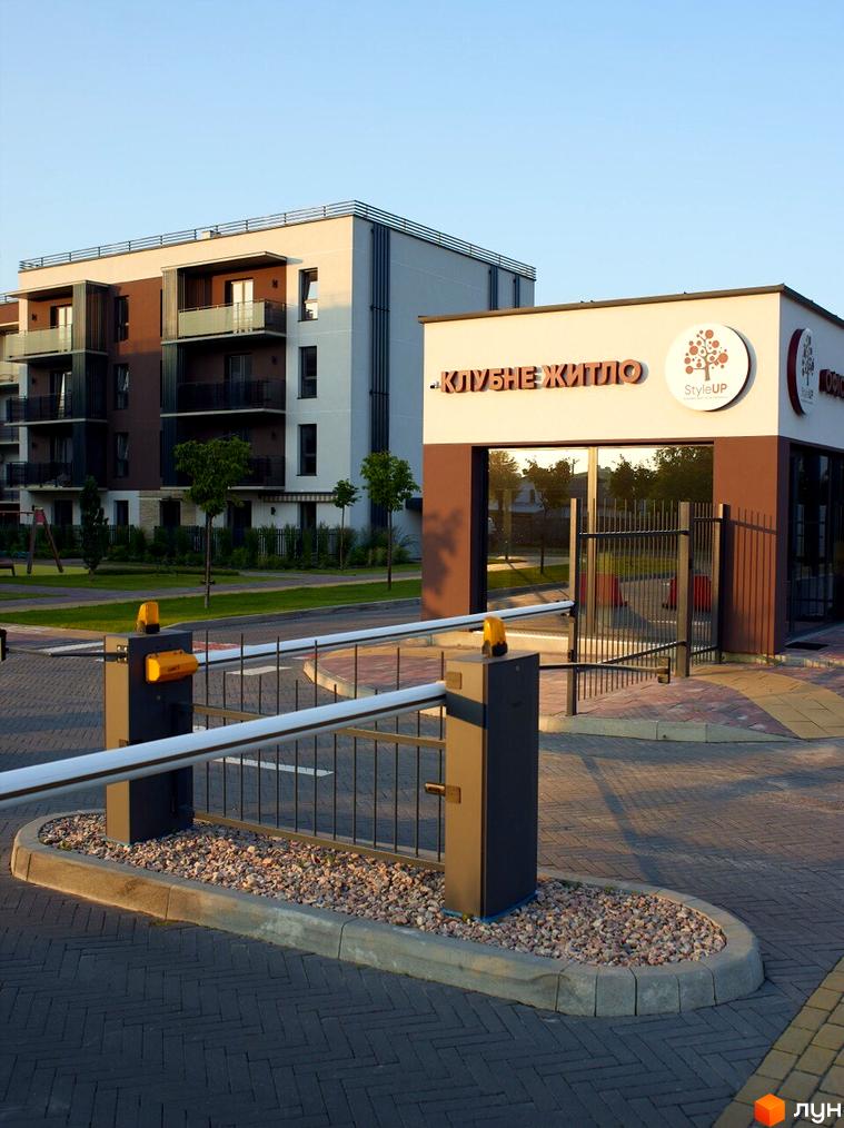 Зображення житлового комплексу Клубне житло StyleUP з фасадом у світлих тонах та коричневими вставками. На передньому плані шлагбаум та доглянута територія.