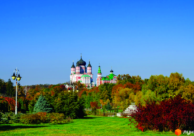 Житловий комплекс Кришталеві джерела на тлі осіннього парку з деревами в жовтих та червоних кольорах. На передньому плані зелений газон та кущі.