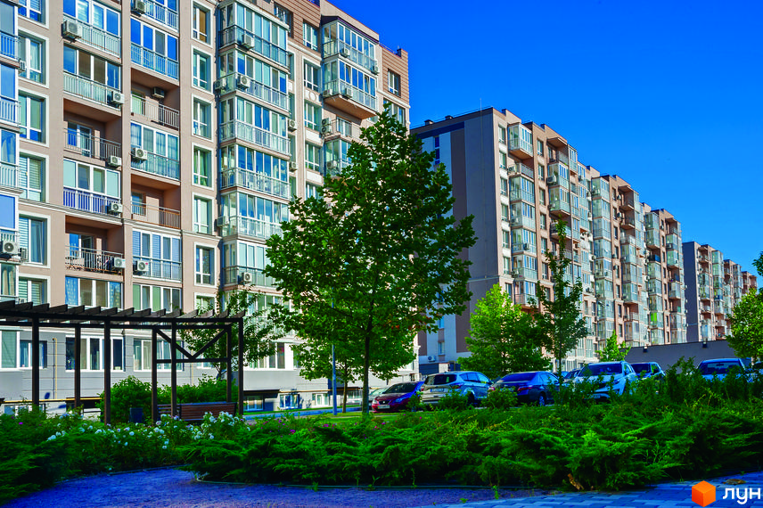 Житловий комплекс Кришталеві джерела. Багатоповерхові будинки з балконами та кондиціонерами. Прибудинкова територія з деревами, кущами та місцями для паркування.