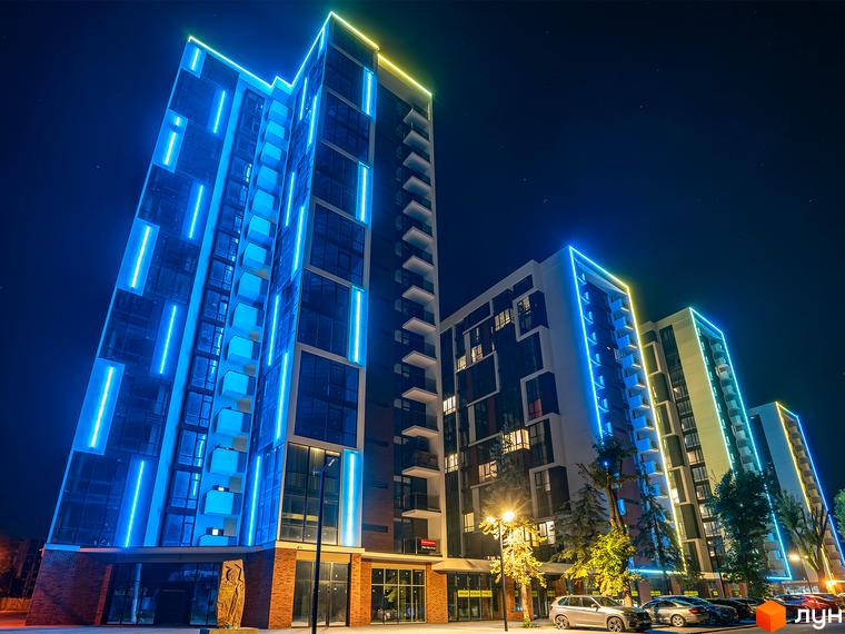 Житловий район Manhattan з яскравою неоновою підсвіткою фасадів у вечірній час. Біля будівель припарковані автомобілі, освітлені ліхтарями.