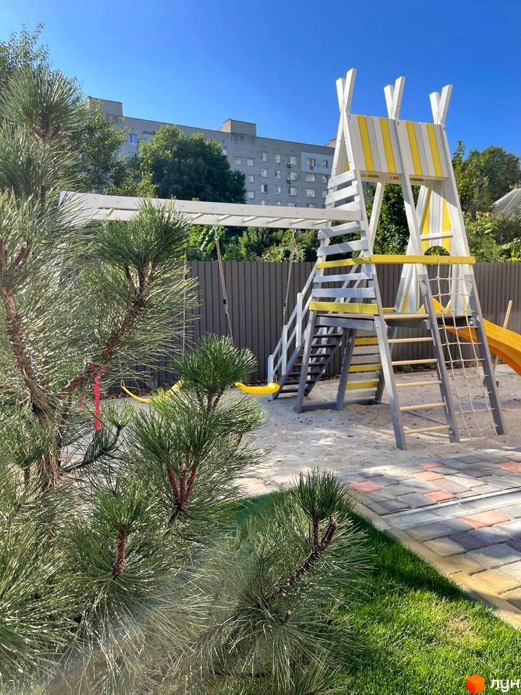 Дитячий майданчик біля житлового комплексу SMART IDEA з гіркою, гойдалкою та пісочницею. Територія облаштована зеленою зоною та покриттям з плитки.