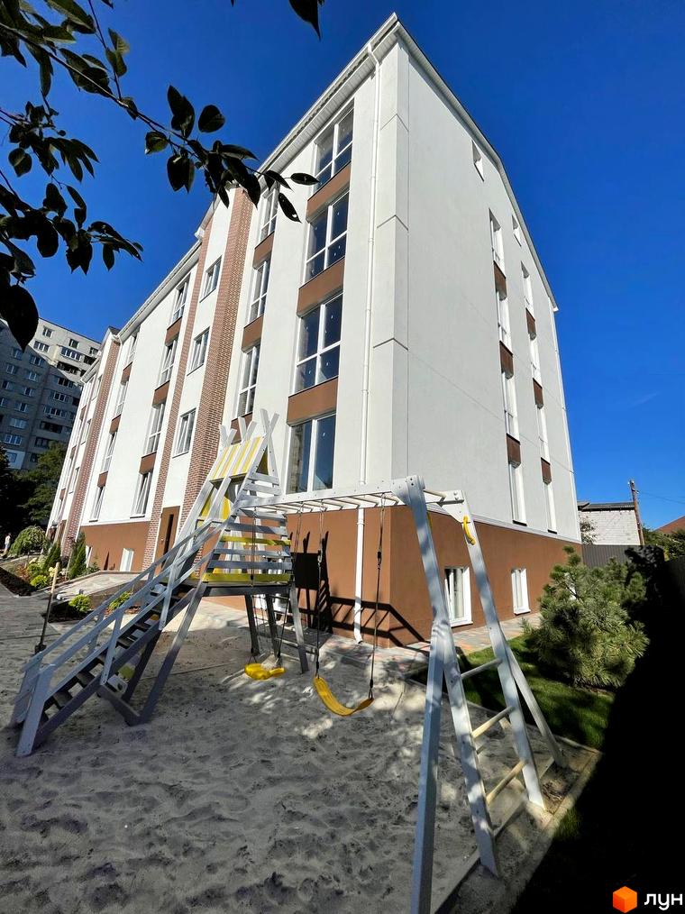 Новобудова ЖК SMART IDEA з біло-коричневим фасадом та дитячим майданчиком з гойдалками на піщаній зоні.