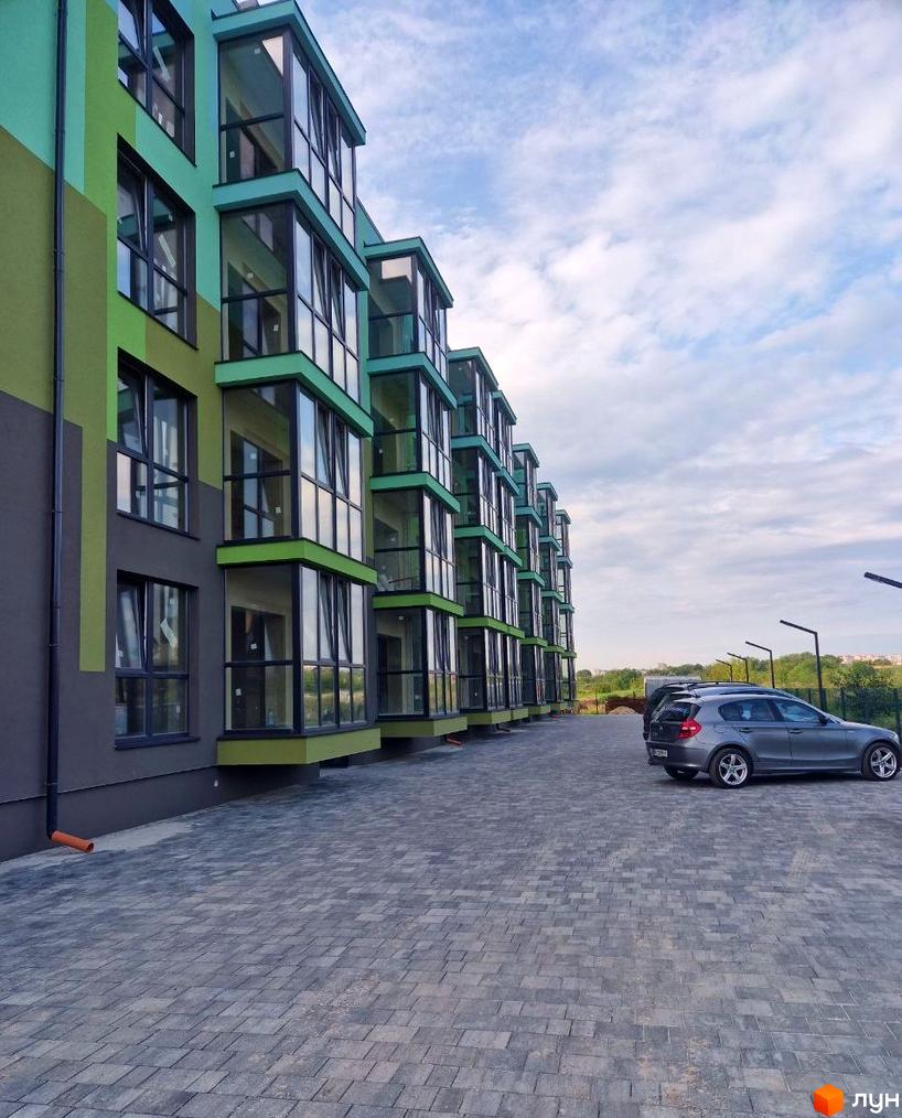 Зовнішній вигляд житлового комплексу Квітковий з зеленими фасадами та скляними балконами, парковка з сірим автомобілем.