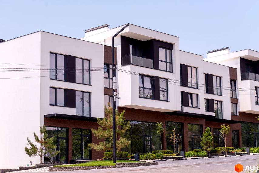 Зовнішній вигляд новобудови ЖК SHERWOOD з білими фасадами, темними вікнами та цегляними елементами. Навколо будівлі розташовані зелені насадження та доглянута прибудинкова територія.