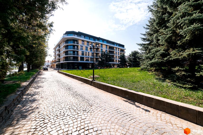 Комплекс RIVER LAND з панорамними вікнами та балконами, розташований на фоні зеленої галявини та дерев. Дорога з бруківки веде до новобудови.