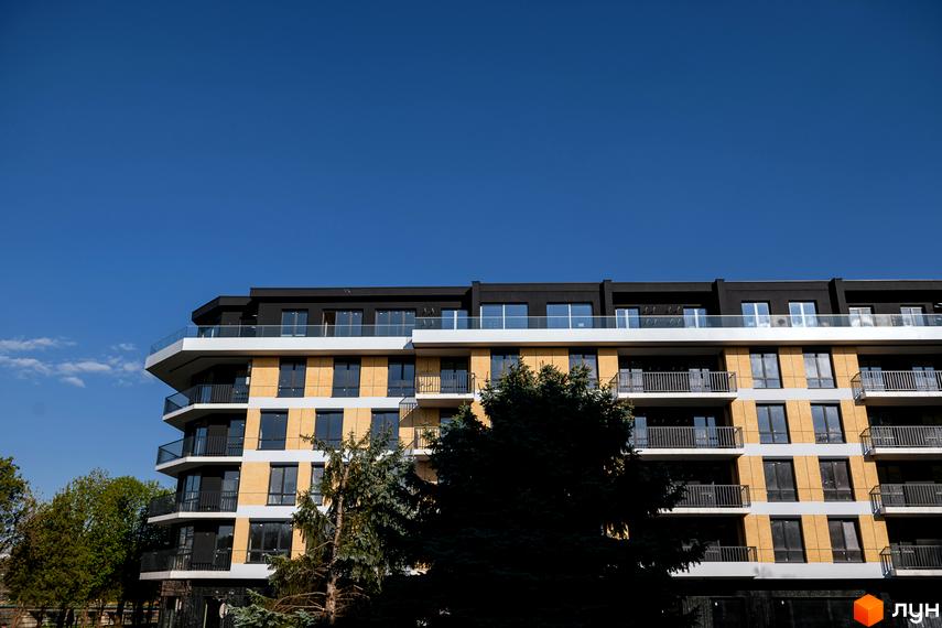 Новобудова ЖК RIVER LAND з жовтим фасадом та скляними балконами на фоні блакитного неба. Будинок має багато вікон та сучасний дизайн.