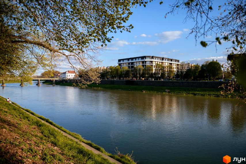 Житловий комплекс RIVER LAND з видом на річку та міст. Сучасна багатоповерхова новобудова з панорамними вікнами та балконами, оточена деревами.