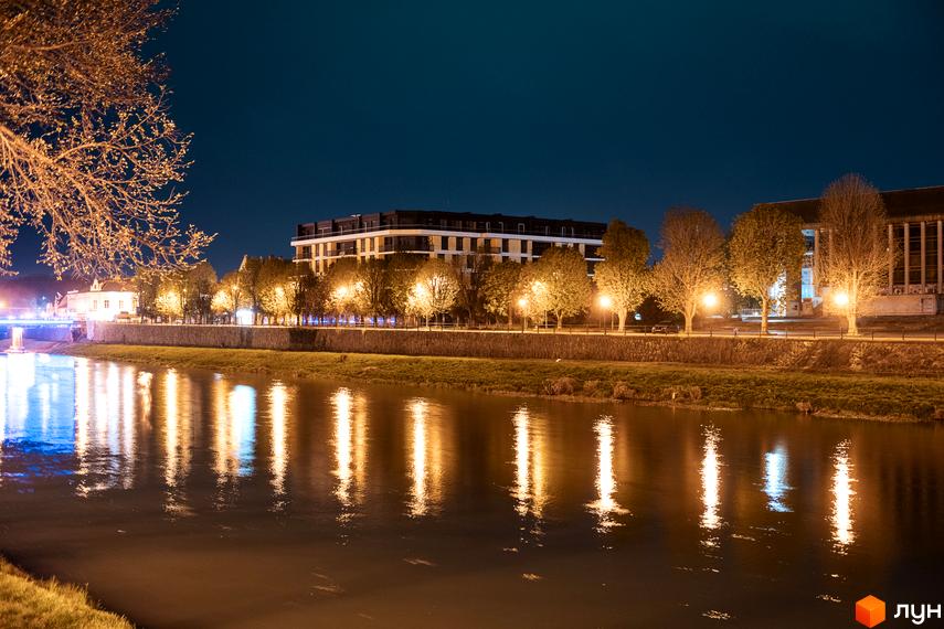 Житловий комплекс RIVER LAND у вечірній час, з відображенням ліхтарів на річці. Сучасна багатоповерхова будівля з балконами та вікнами.