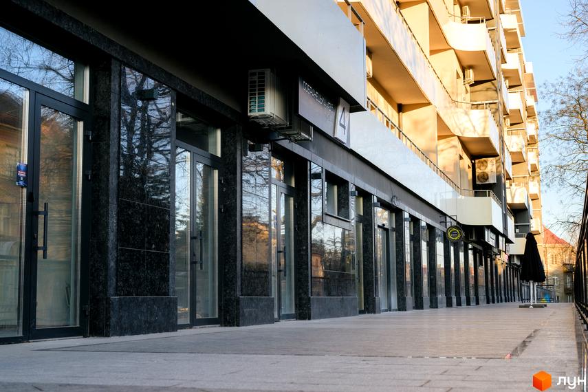 Зовнішній вигляд житлового комплексу DREAM CITY з темним оздобленням фасаду та скляними вітринами. Поруч розташована доглянута прибудинкова територія з бруківкою та деревами.