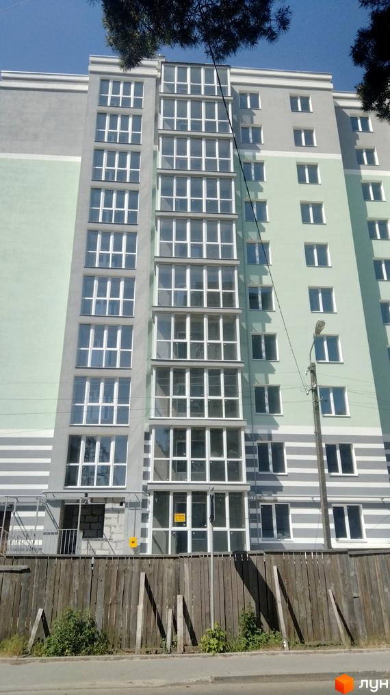 Зовнішній вигляд новобудови на вул. Волковича, 25, з великою кількістю вікон та засклених балконів, на тлі блакитного неба. Перед будинком розташований дерев'яний паркан.