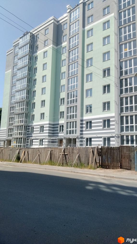 Багатоповерховий житловий комплекс з панорамними вікнами та балконами, оздоблений світло-зеленими та сірими фасадами, з дерев'яною огорожею та дорогою перед будинком.