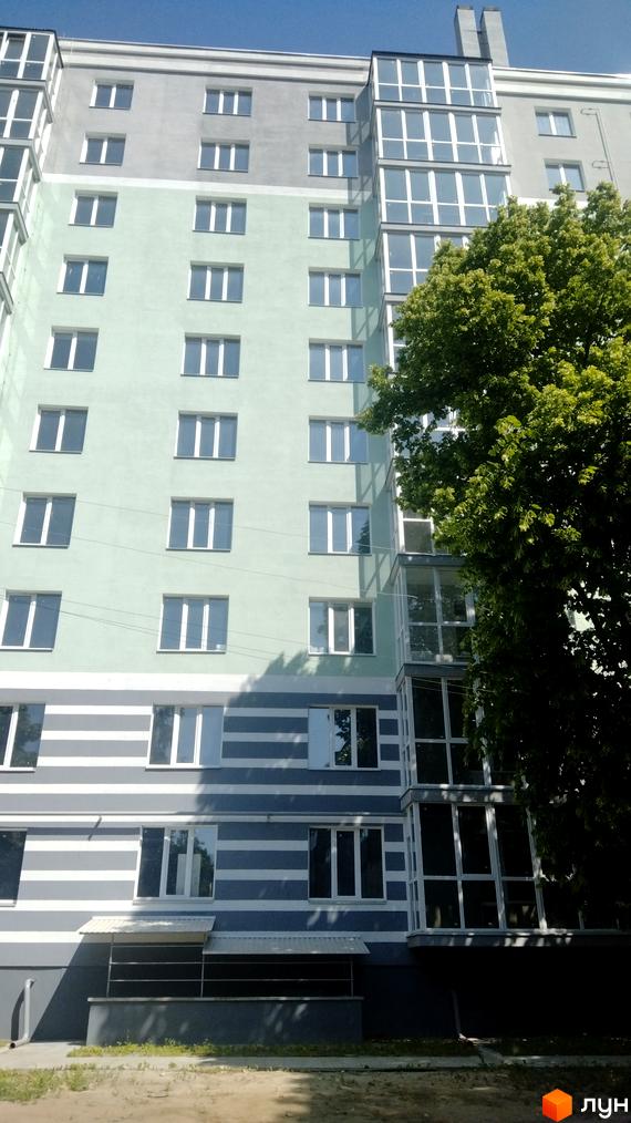 Фасад житлового комплексу на вул. Волковича, 25, з світло-зеленими та сірими секціями, великими вікнами та скляним балконом. Поруч росте зелене дерево.