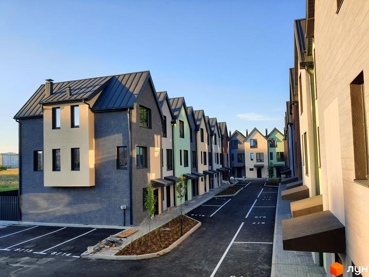 Ряд різнокольорових таунхаусів New Smart 11 з темними дахами та парковкою перед ними. Зовнішній вигляд будинків та прибудинкова територія.