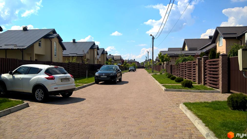 Котеджне містечко Наше містечко. Будинки з темними дахами та світлими стінами, розташовані вздовж вулиці з бруківки, з доглянутими газонами та парканами.