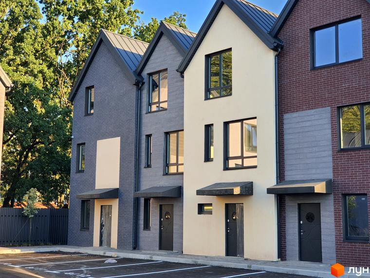Таунхауси New Smart 11 – це двоповерхові будинки з сірої та кремової цегли з темними дахами та вікнами. Перед будинками розташована парковка з розміткою.