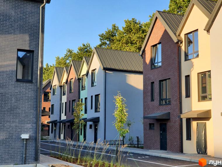 Таунхауси New Smart 11 – це низка двоповерхових будинків з цегляним фасадом та темним дахом, розташованих вздовж дороги з зеленими насадженнями.