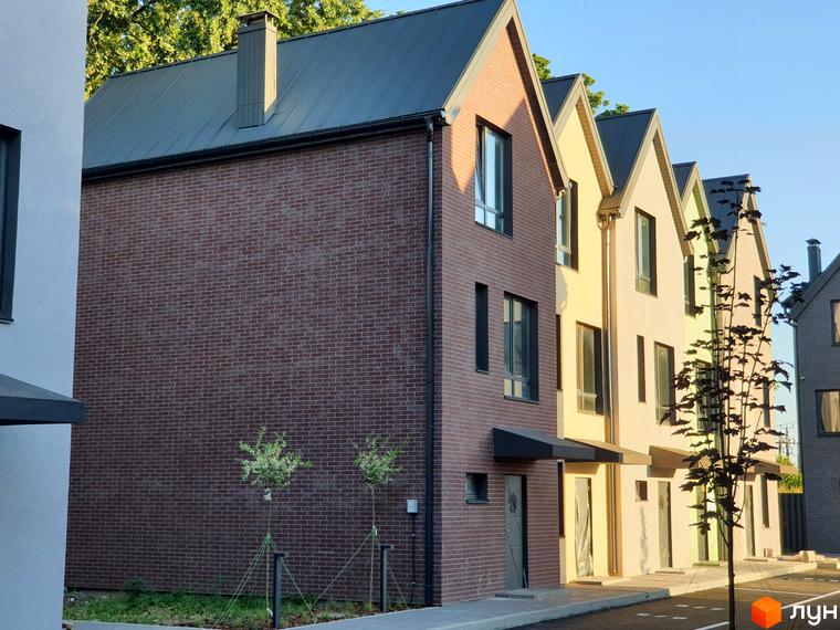 Таунхауси New Smart 11 - це комплекс з цегляних будинків з темними дахами та вікнами, розташований на тлі блакитного неба. Перед будинками облаштована прибудинкова територія з доріжками та зеленими насадженнями.