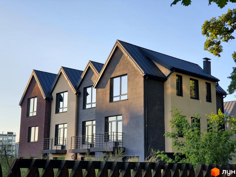 Таунхауси New Smart 11 – це триповерхові будинки з темними дахами та фасадами різних кольорів, розташовані на фоні блакитного неба. Навколо будівель видно зелені насадження та дерев'яний паркан.