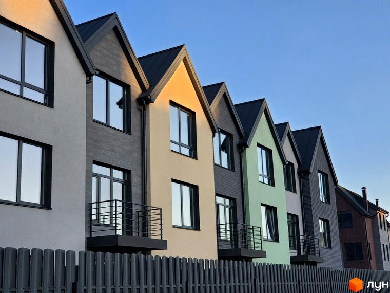 Ряди сучасних таунхаусів New Smart 11 з різнокольоровими фасадами та чорними дахами на тлі блакитного неба. Перед будинками встановчено сірий паркан.