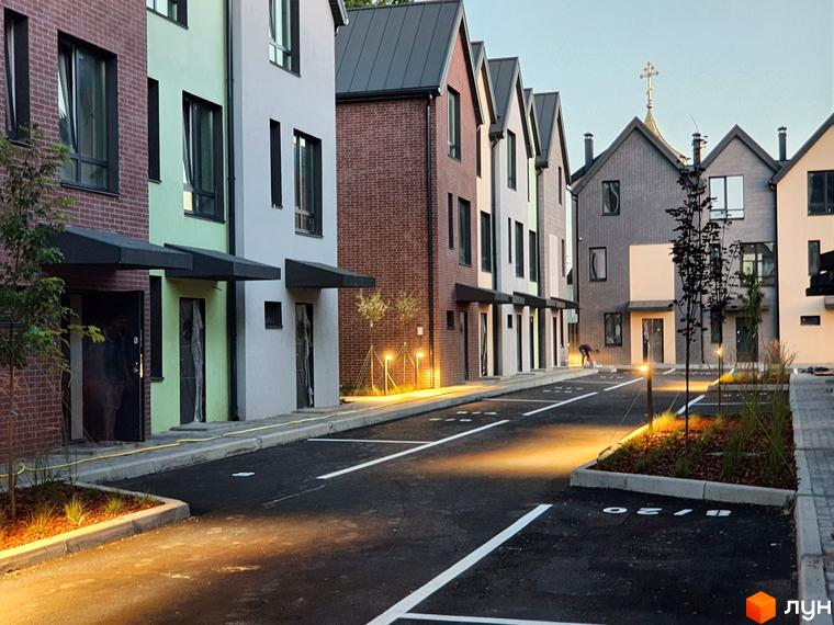 Таунхауси New Smart 11 з фасадами різних кольорів та парковкою з розміткою. Навколо новобудови висаджені молоді дерева та освітлення.