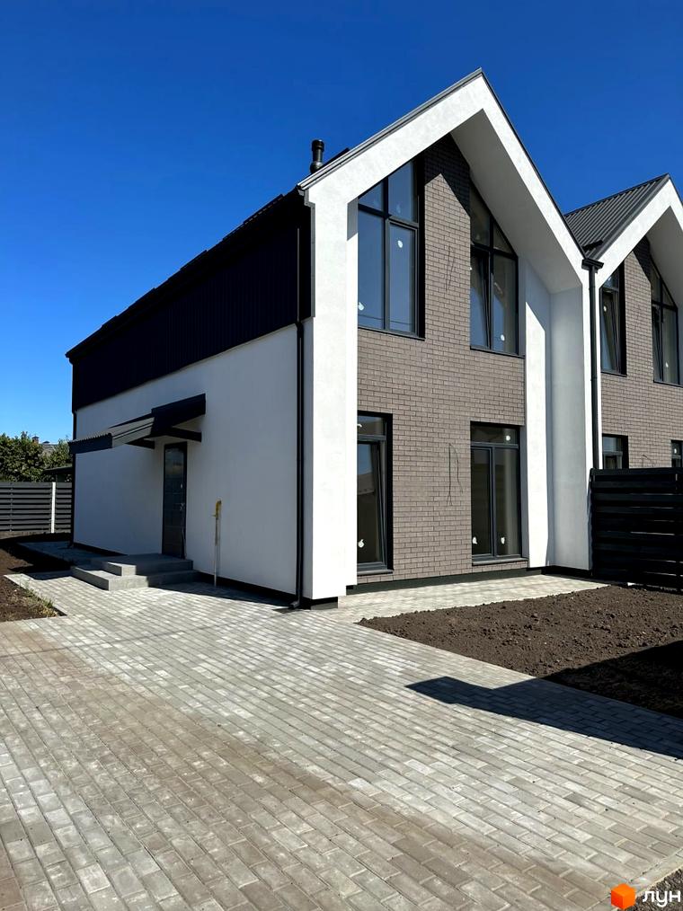 Зовнішній вигляд котеджного містечка КМ New Village: білі фасади з темними елементами, цегляні вставки та панорамні вікна. Прибудинкова територія з бруківкою та зеленими насадженнями.