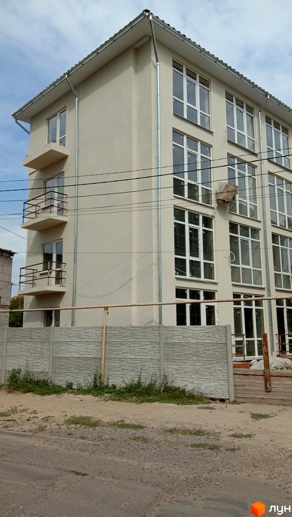 Будівельний проєкт Першотравневий: світлий багатоповерховий будинок з балконами та великими вікнами на тлі блакитного неба. Попереду видно бетонний паркан та зелень.