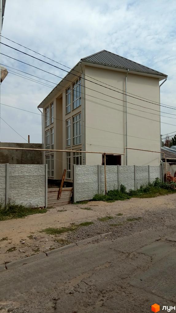 Зовнішній вигляд триповерхової новобудови ЖК Першотравневий з великими вікнами та сірим дахом. Територія огороджена бетонним парканом.