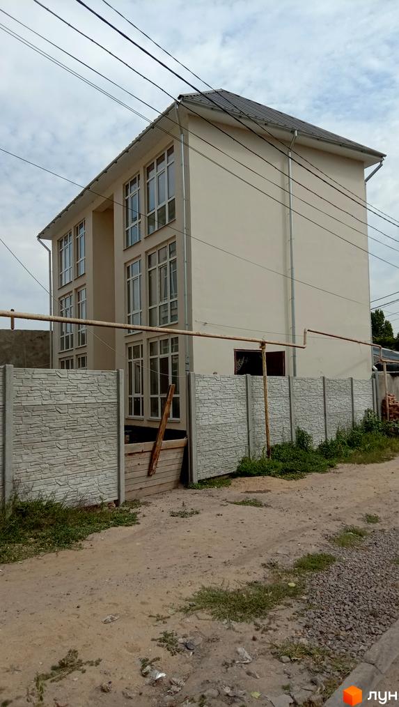 Зовнішній вигляд новобудови ЖК Першотравневий з великими вікнами та сірим дахом. Навколо будинку розташована бетонна огорожа та зелені насадження.