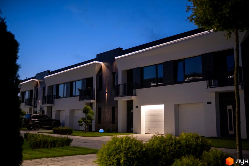 Котеджне містечко КМ SHERWOOD: фасади будинків з підсвічуванням та гаражними воротами, прибудинкова територія з газоном та кущами.