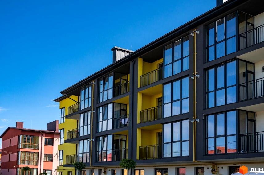 Фасад житлового комплексу Італійський з жовтими та сірими елементами, великими вікнами та балконами на тлі блакитного неба. Поруч розташована червоно-помаранчева будівля.