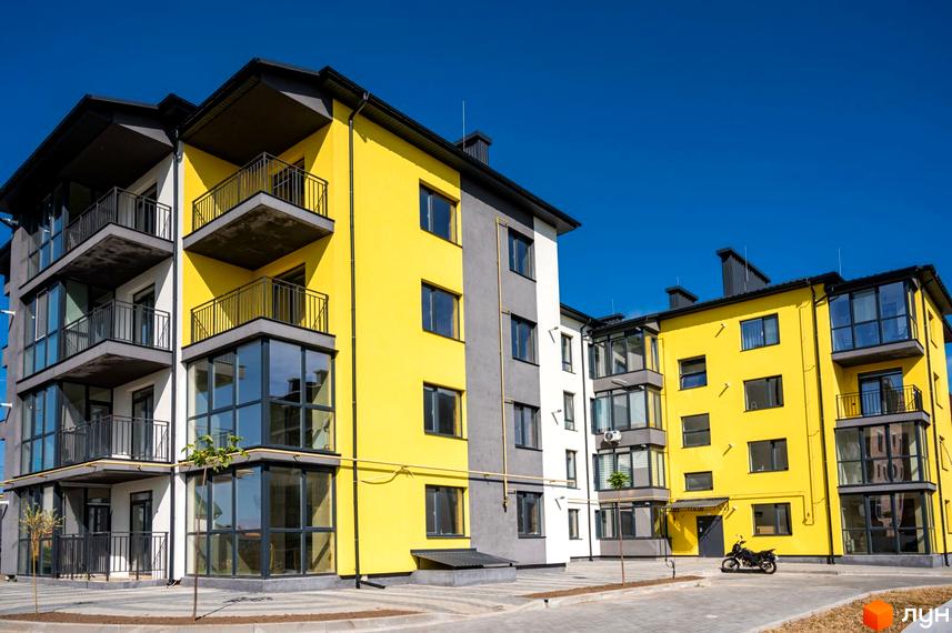 Фасад житлового комплексу Італійський у жовто-сірих тонах з балконами та великими вікнами. На прибудинковій території видно бруківку та мотоцикл.