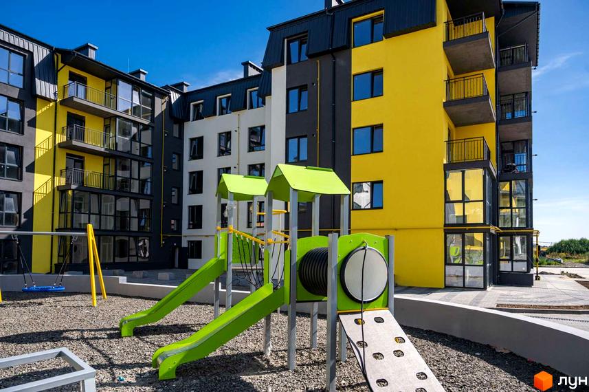 Житловий комплекс Італійський з яскравою жовтою фасадною частиною та дитячим майданчиком із зеленими гірками та гойдалками.