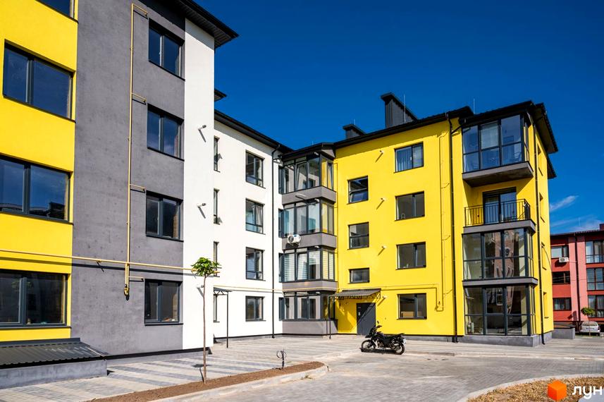 Житловий комплекс Італійський з яскравими жовтими та сірими фасадами, скляними балконами та вікнами. Територія з бруківкою, деревом та мотоциклом.