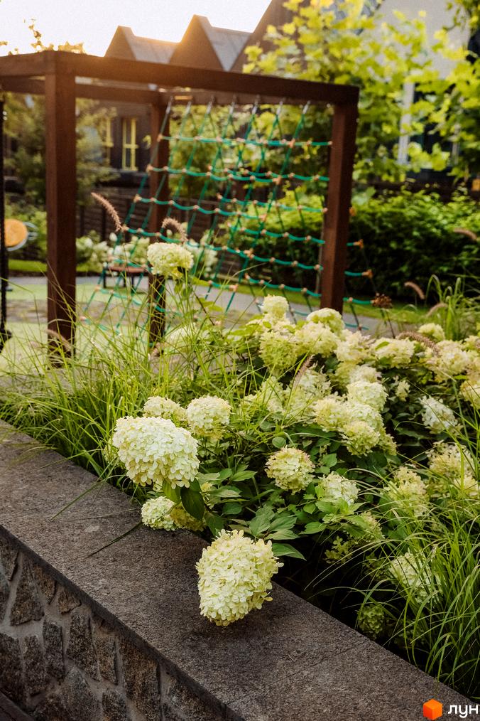 Котеджне містечко Oak Grove Town: будинок з темним дахом, клумба з білими гортензіями, травою, кам'яною огорожею та дитячий майданчик.