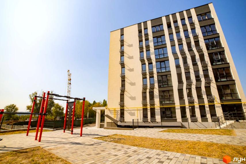 Житловий комплекс Сонячний: багатоповерховий будинок зі світлим фасадом, темними вертикальними вставками та вікнами. На прибудинковій території розташований спортивний майданчик.