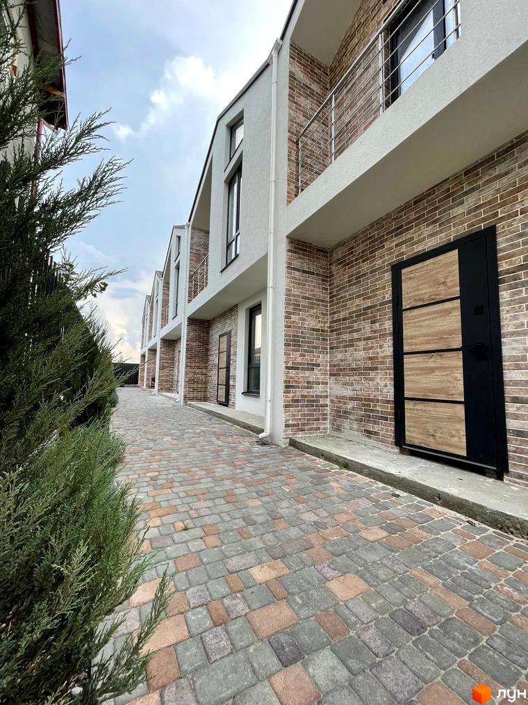 Таунхауси West Homes з цегляним фасадом та дерев'яними вставками на дверях. Перед будинками вимощена бруківкою доріжка та зелені насадження.