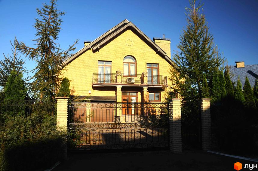Котеджне містечко КМ Вишневе містечко. Фасад жовтої цегляної двоповерхової будівлі з балконами та вікнами. Навколо будинку зелені дерева та огороджена територія.
