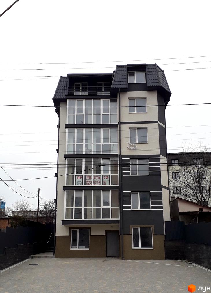 Багатоповерхова новобудова з панорамними вікнами та сірими елементами фасаду, розташована на вимощеній бруківкою території.