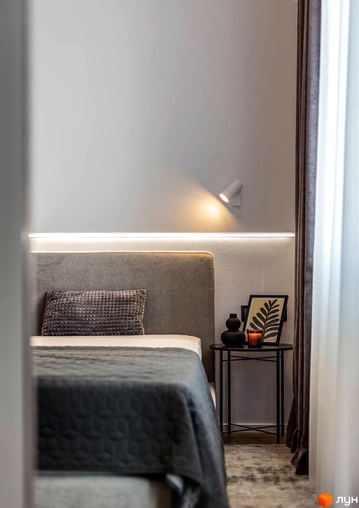 Затишна спальня в ЖК Nebo з великим узголів'ям та декоративною подушкою. Поруч столик з вазою, свічкою та картиною.