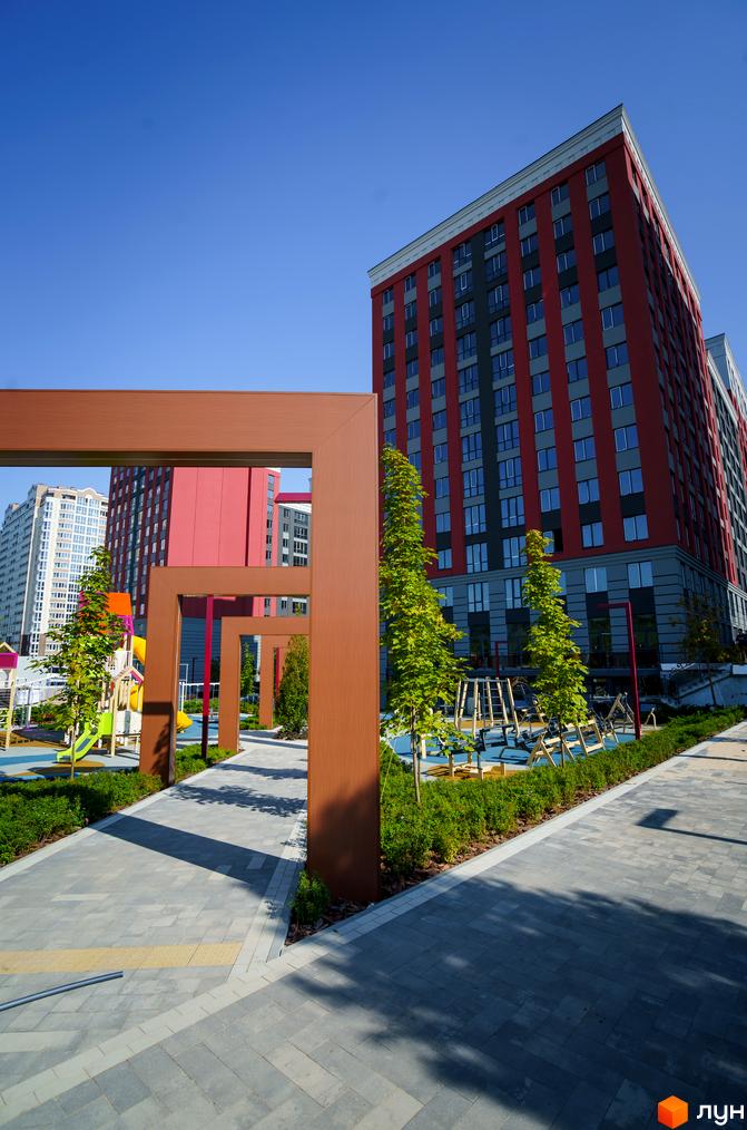 Житловий комплекс Irpin City: червоно-сіра новобудова з великою кількістю вікон. Поруч облаштований дитячий майданчик та зелена зона з деревами.