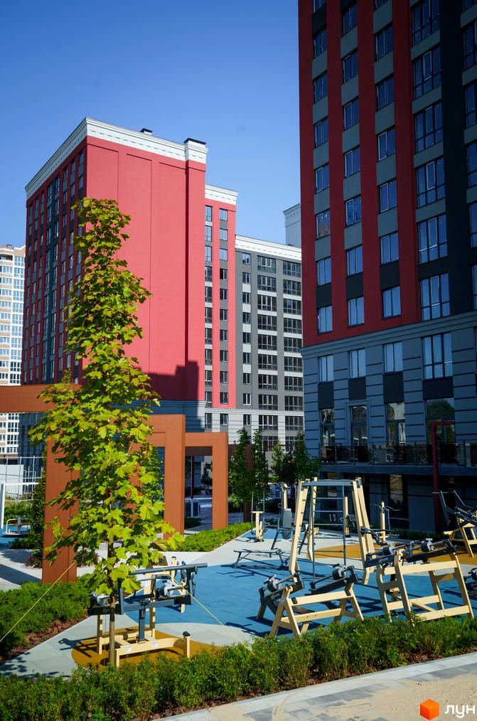 Житловий комплекс Irpin City з червоними та сірими багатоповерховими будинками. На прибудинковій території розташований спортивний майданчик з тренажерами та зелені насадження.