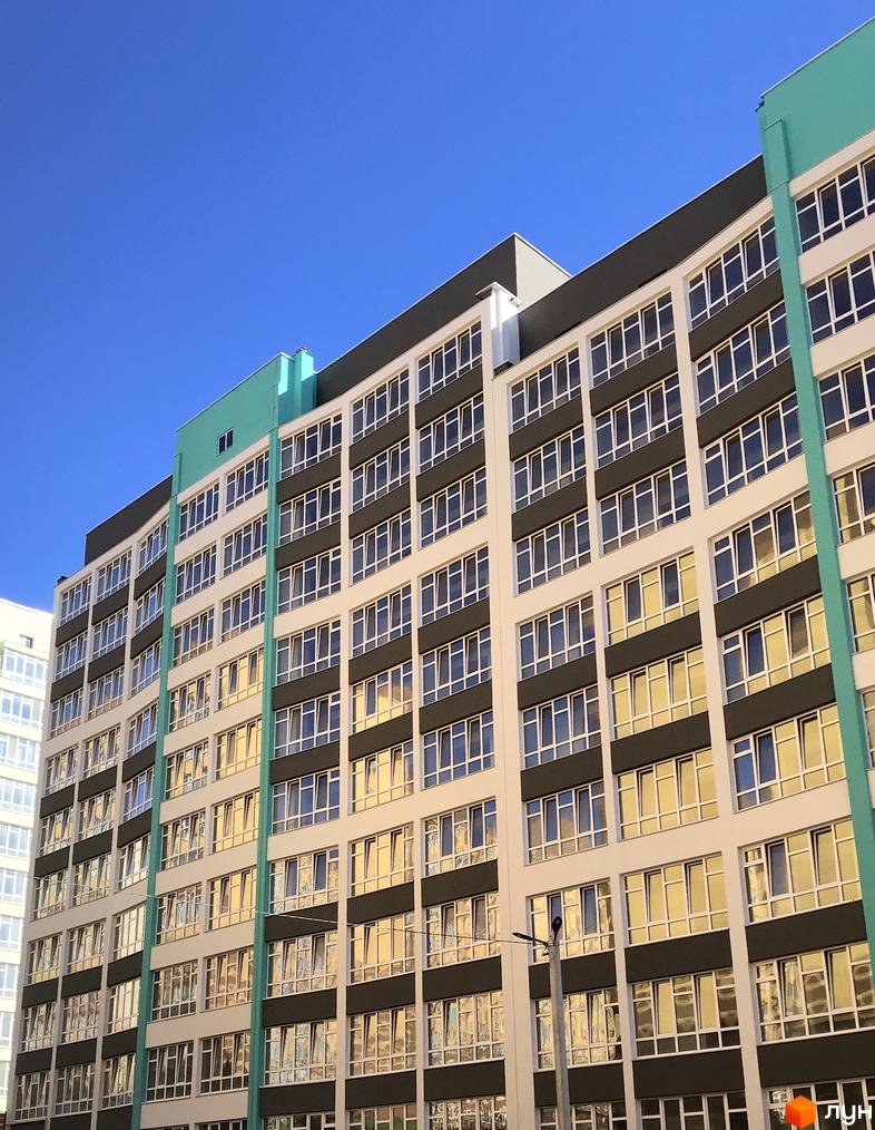 Зовнішній вигляд ЖК Новобудова: багатоповерховий будинок з великою кількістю вікон. Фасад світлий з темними вставками та бірюзовими елементами.