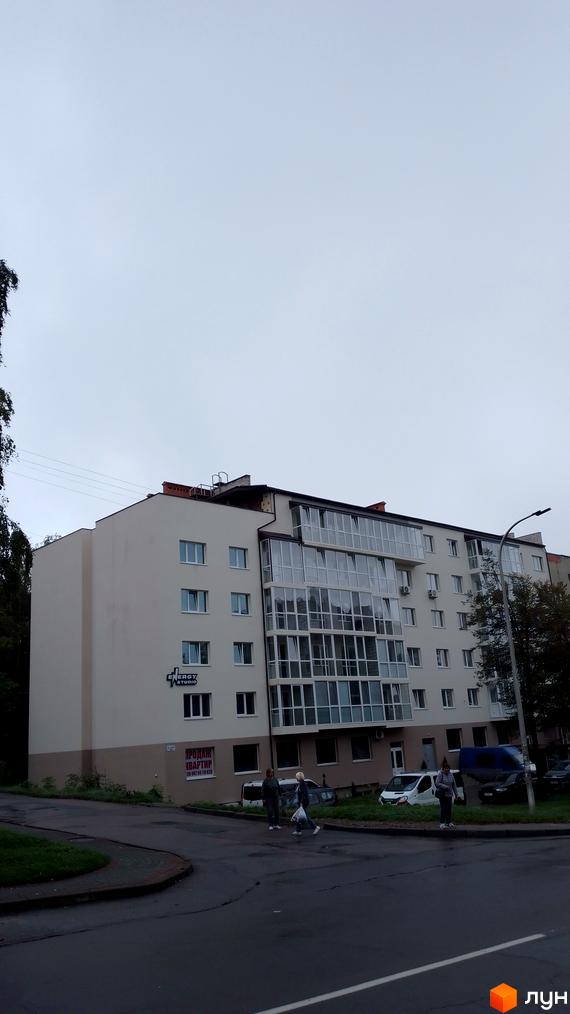 Фасад багатоповерхової новобудови з панорамними вікнами та балконами. Поруч дорога та зелена зона з деревами.