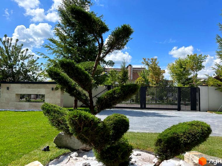 КМ Riviera City Park: зелений газон, доглянуті кущі та сучасні ворота перед будинком. Навколо багато зелені та блакитне небо з хмарами.